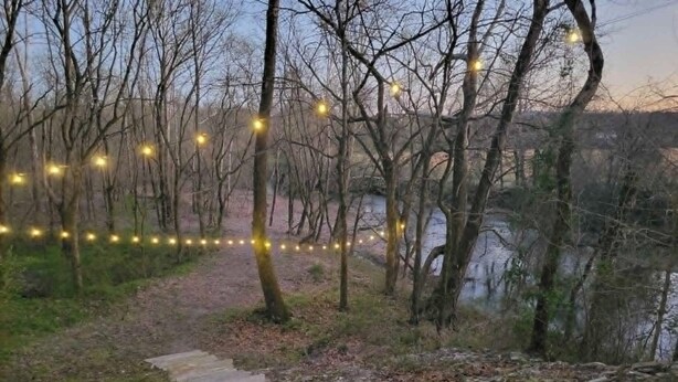 Ein Pfad mit Steinstufen führt durch einen bewaldeten Bereich mit kahlen Bäumen, gesäumt von einer Lichterkette.