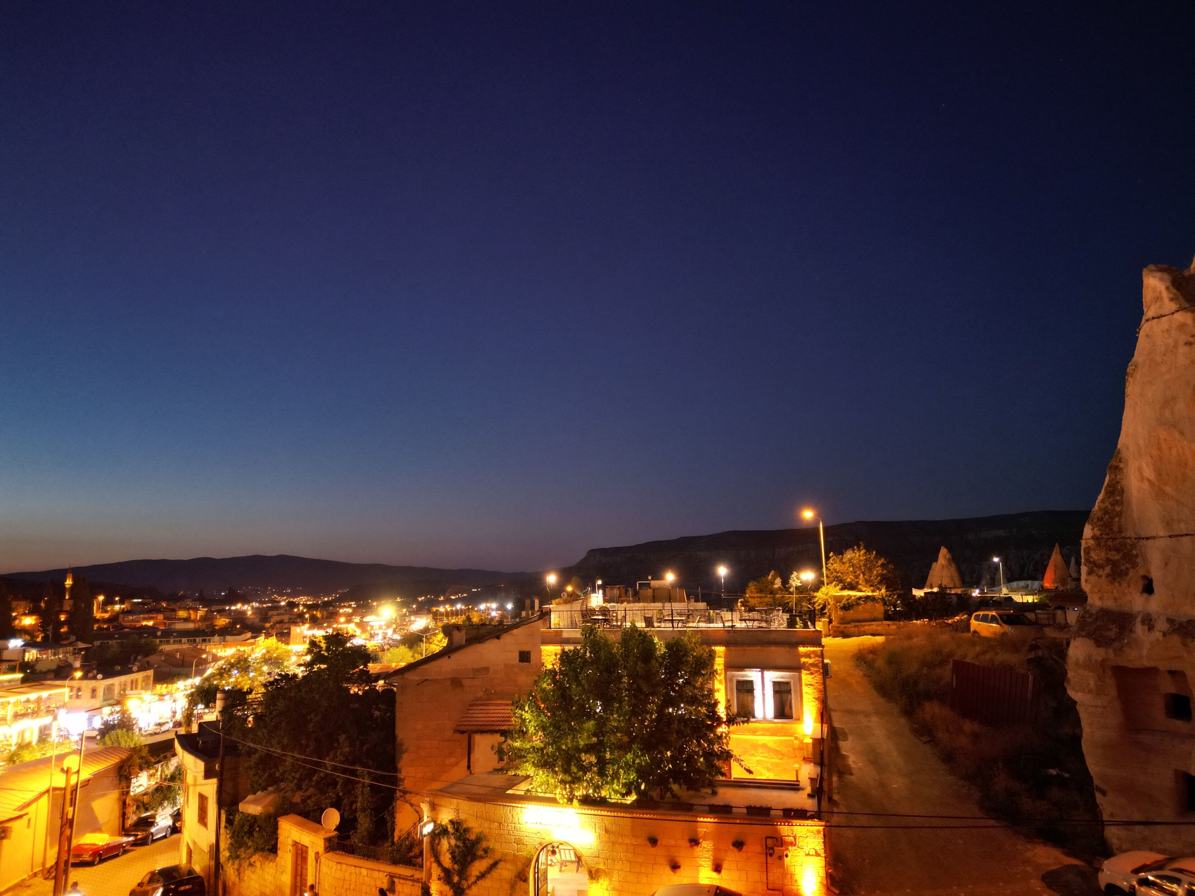 A nighttime view of a town with illuminated streets and buildings, a prominent hill in the background, and a large rock formation on the right.