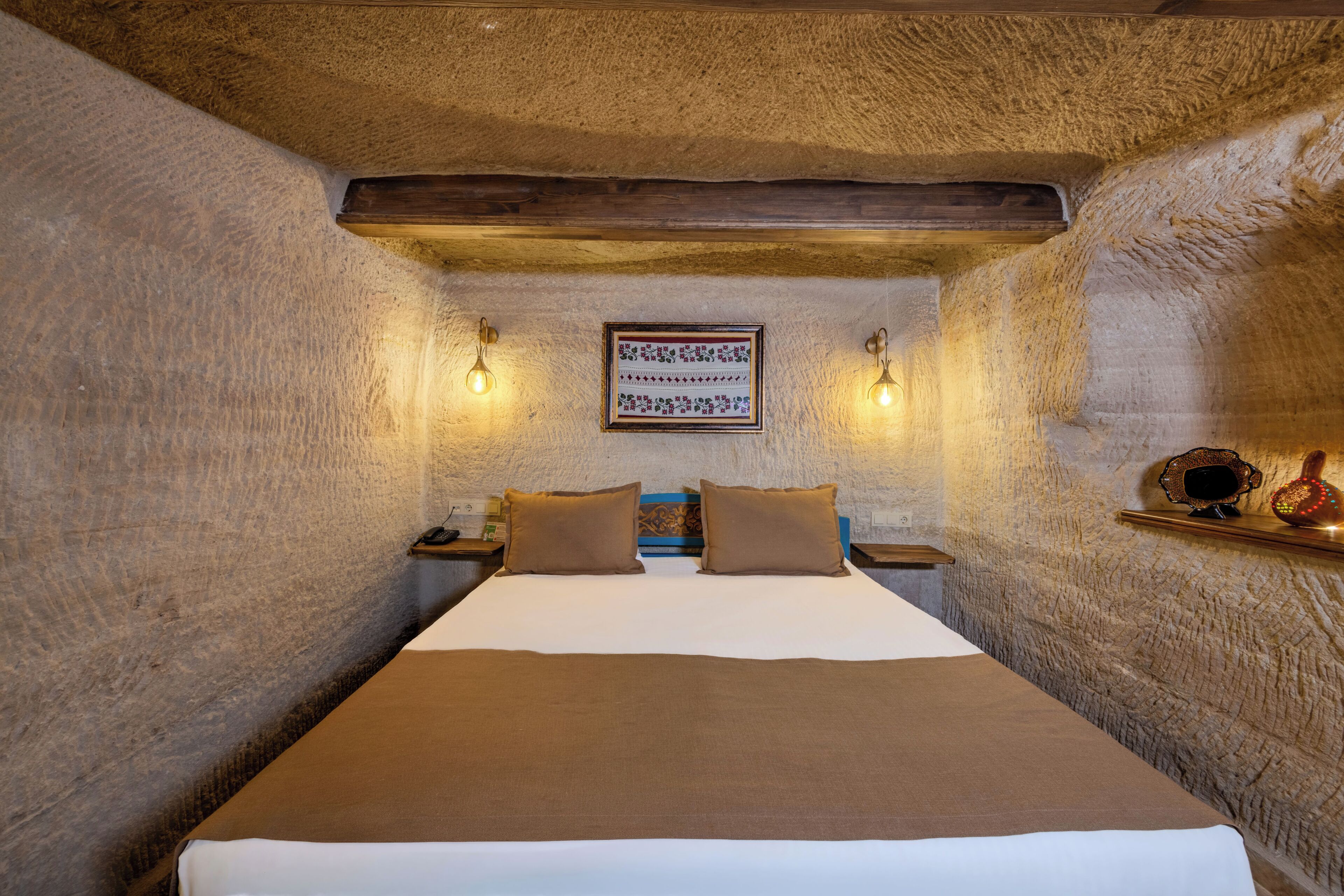A room with a bed, two wall-mounted light fixtures, a framed picture, and a shelf with decorative items.