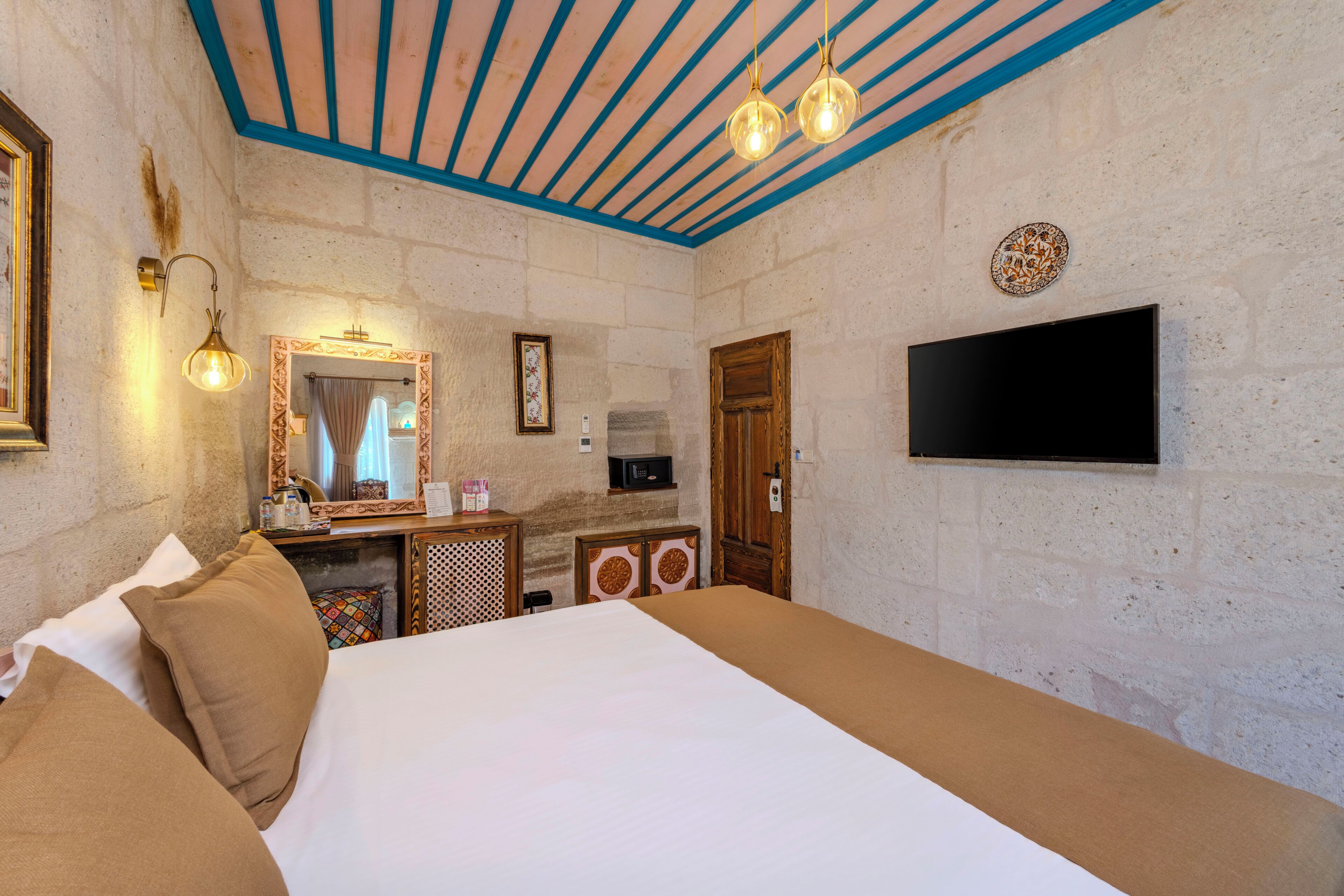 Habitación de hotel con cama, cómoda, espejo, televisor y techo con vigas azules.