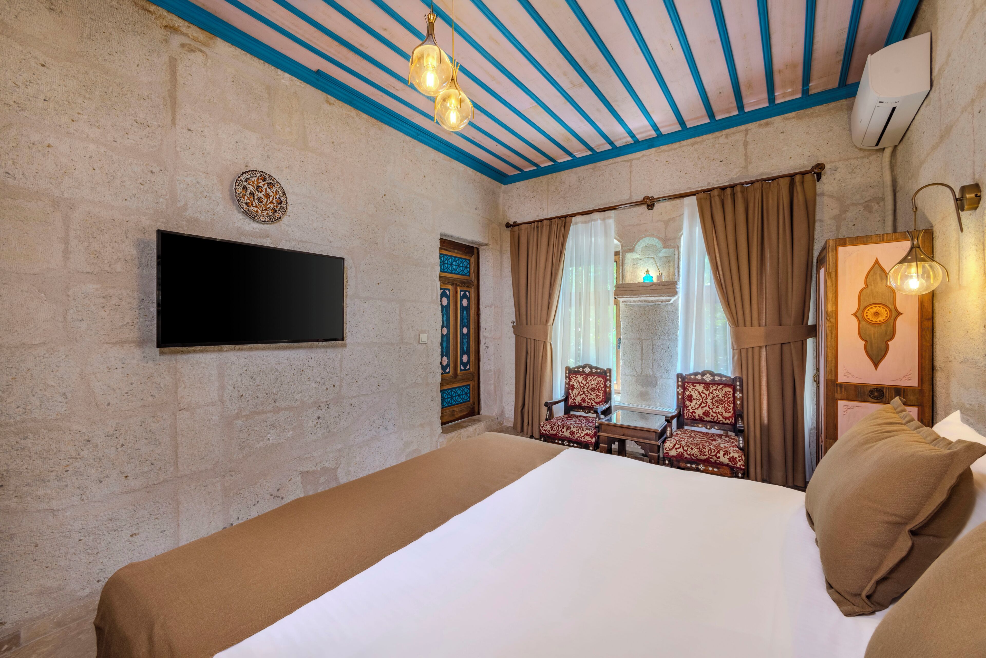 A hotel room with a bed, a flat-screen TV mounted on the wall, a window with curtains, a chair, and a wall-mounted lamp.