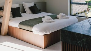 Une chambre avec un plafond en pente, une tête de lit en bois, un lit recouvert de draps blancs, un plaid vert et une vue sur l’extérieur par une fenêtre.