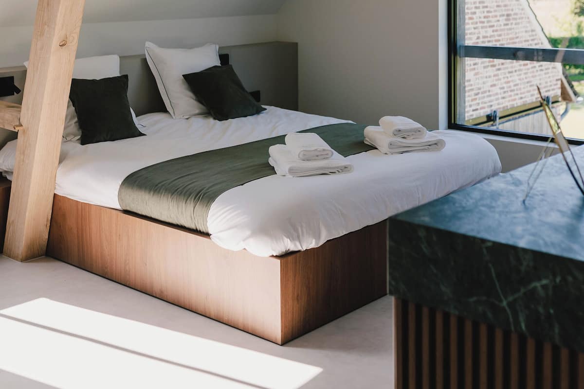 A bedroom with a sloped ceiling, a wooden headboard, a bed with white linens, a green throw, and a view of the outside through a window.