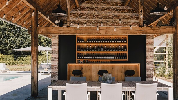 Un espace bar extérieur avec un mur en briques, un plafond en bois et une longue table en bois entourée de chaises blanches.