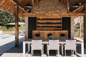 An outdoor bar area with a brick wall, wooden ceiling, and a long wooden table with white chairs. - B&B Entre Nous (Herk-de-Stad)