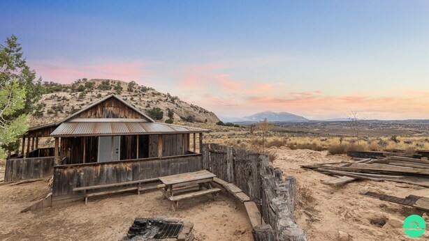 A rustic wooden building with a metal roof, surrounded by a wooden fence and picnic tables, set against a desert landscape with distant mountains. - The Sunset Retreat Cabin (Monticello)