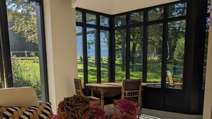 Un saló modern amb un sofà, una taula de centre de fusta amb un ram de flors, i una zona de menjador amb taula i cadirees. Grans finestres ofereixen vista a un jardí.