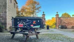 Un entorno rústico al aire libre con una mesa y bancos de madera, un edificio de piedra y una estructura decorativa con motivos de oso y hongo.