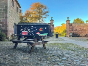 Un entorno rústico al aire libre con una mesa y bancos de madera, un edificio de piedra y una estructura decorativa con motivos de oso y hongo.