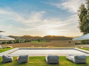 Un espace piscine avec vue sur des collines verdoyantes et un coucher de soleil. On y trouve trois fauteuils en forme de sac et une chaise longue.