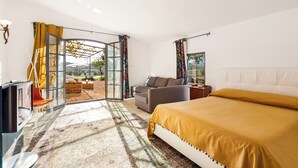 A modern bedroom with a large bed, a sofa, a TV, and a glass door leading to an outdoor area. - Casale Rondinella 6+2, Emma Villas (Manciano)