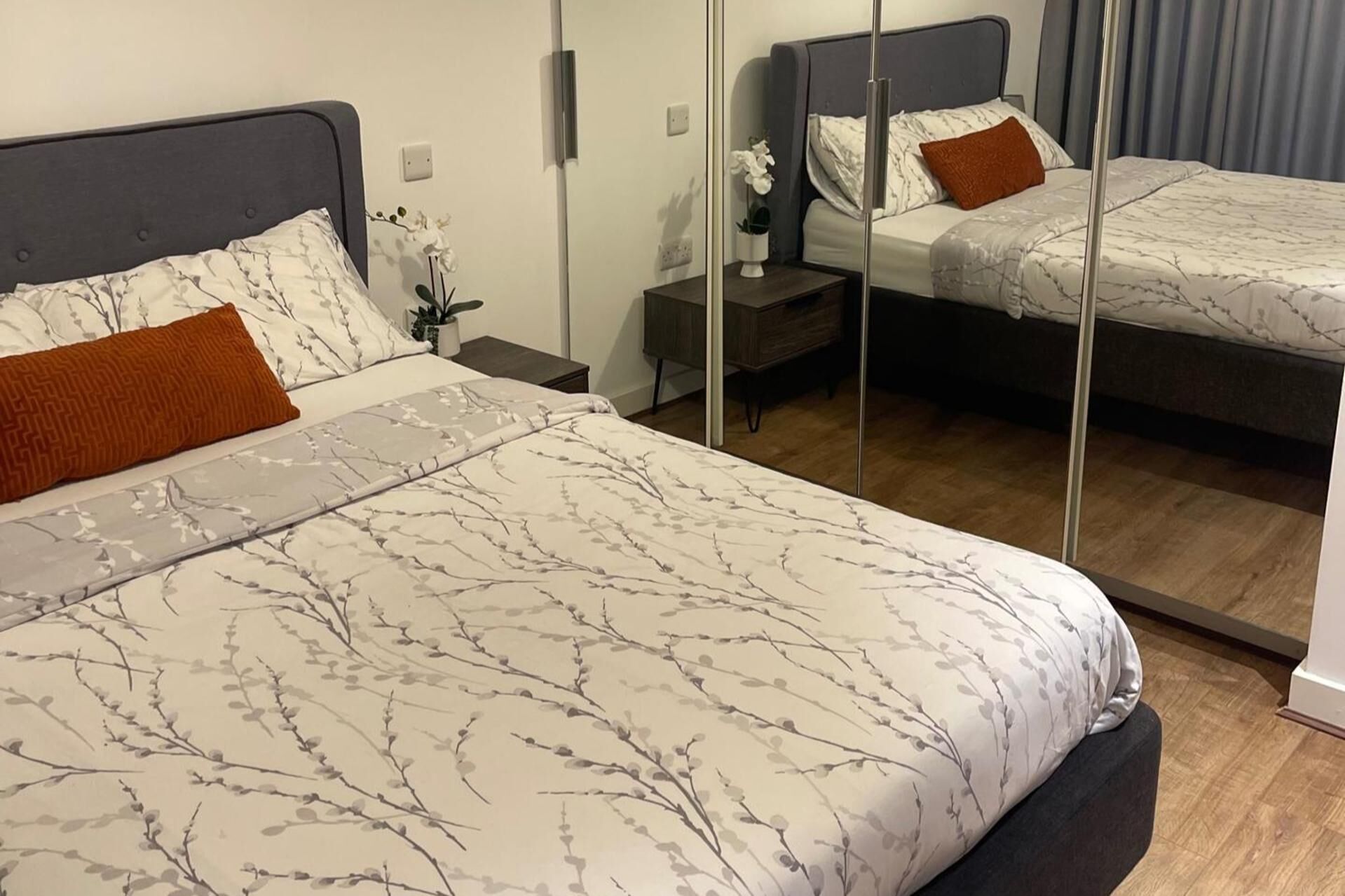 A hotel room with two beds, each with a grey headboard and white bedding with a leaf pattern. There is a small bedside table with a plant and a mirror on the wall.