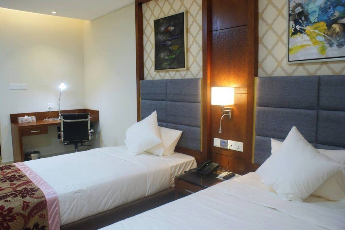 A hotel room with two beds, a desk, a chair, a lamp, and artwork on the walls.