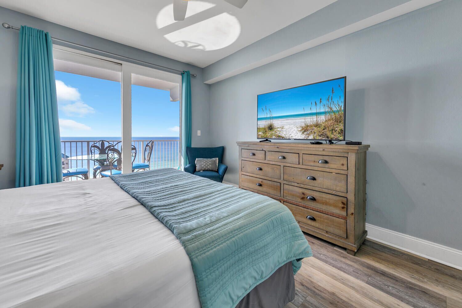 A bedroom with a large bed, a wooden dresser, a television on top, and a balcony with a view of the beach.