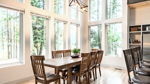 Comedor con mesa y sillas de madera, ventanas amplias con vista al bosque y una lámpara colgante.