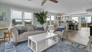 Ein geräumiges Wohnzimmer mit einem beigen Sofa, einem weißen Couchtisch und einem Deckenventilator. Ein großes Fenster bietet einen Blick ins Freie.