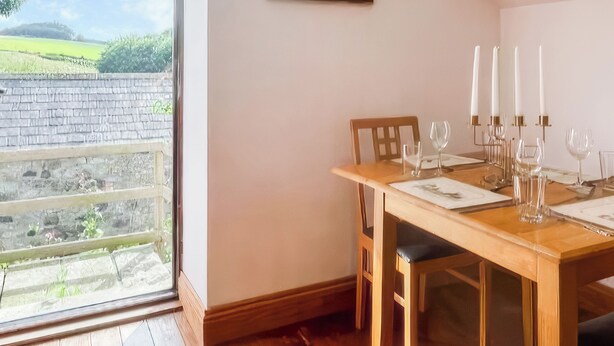 A dining area with a wooden table set for six, a window with a view of a brick wall and greenery, and a wooden floor.