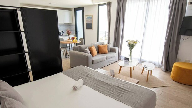 Habitación de hotel moderna con una cama grande, un sofá, una mesa de comedor y una zona de cocina al fondo.