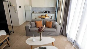 Una sala moderna con un sofá gris, dos mesas blancas y un florero con flores. Se ve una cocina al fondo, con armarios blancos y una mesa de comedor.