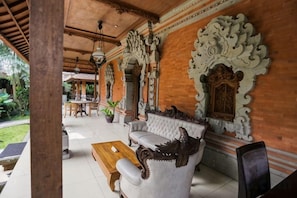 Un espacio de estar al aire libre techado, con un sofá y una mesa de madera, rodeado de decoraciones ornamentales en las paredes y una planta en maceta.