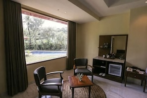 A hotel room with a large window, a desk, two chairs, a mirror, and a small table with a decorative item. - Ubud Hotel & Cottages (Sukun)