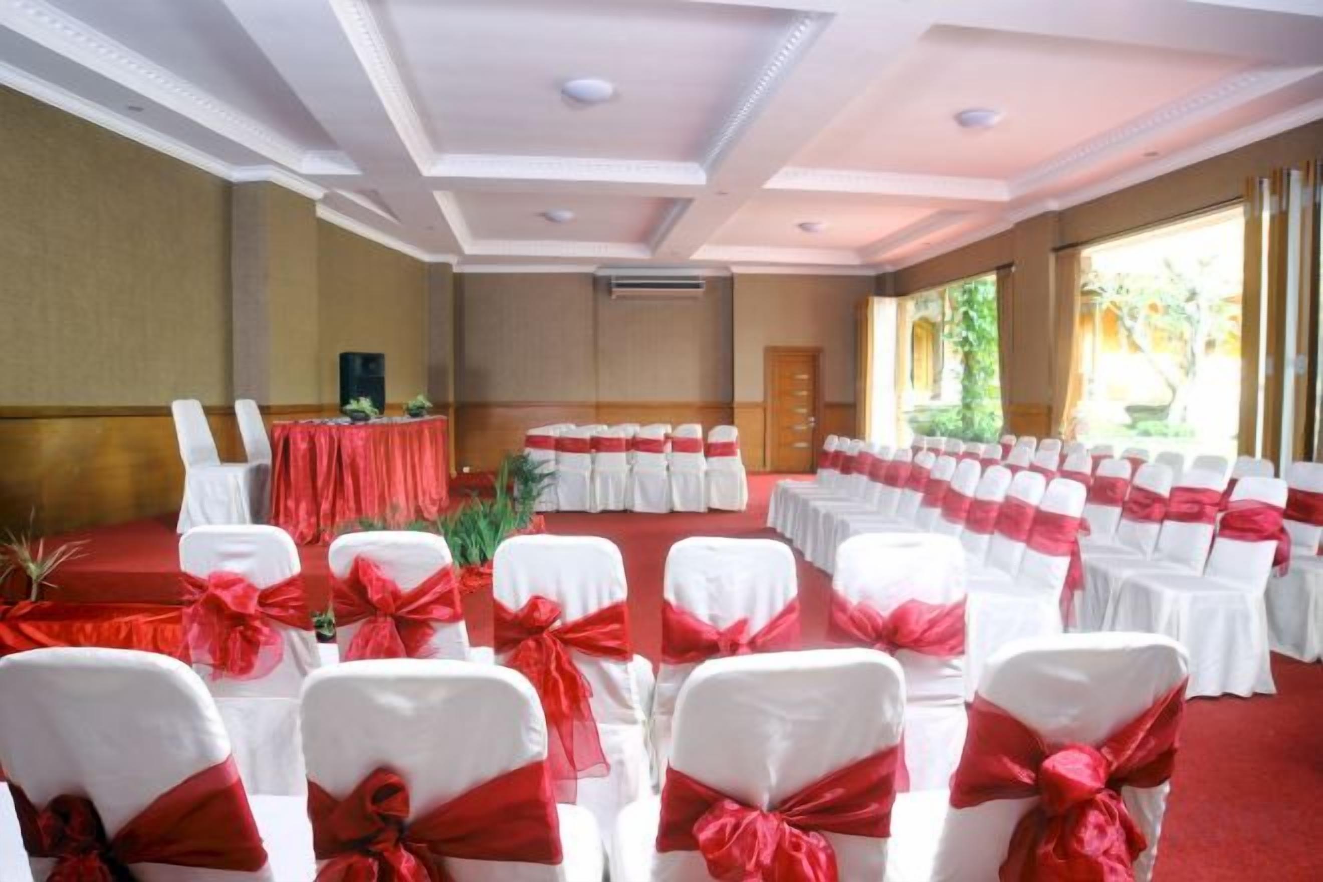 Uma sala de conferências com fileiras de cadeiras brancas com fitas vermelhas, um palco com toalha de mesa vermelha e vista para o verde pelas janelas.