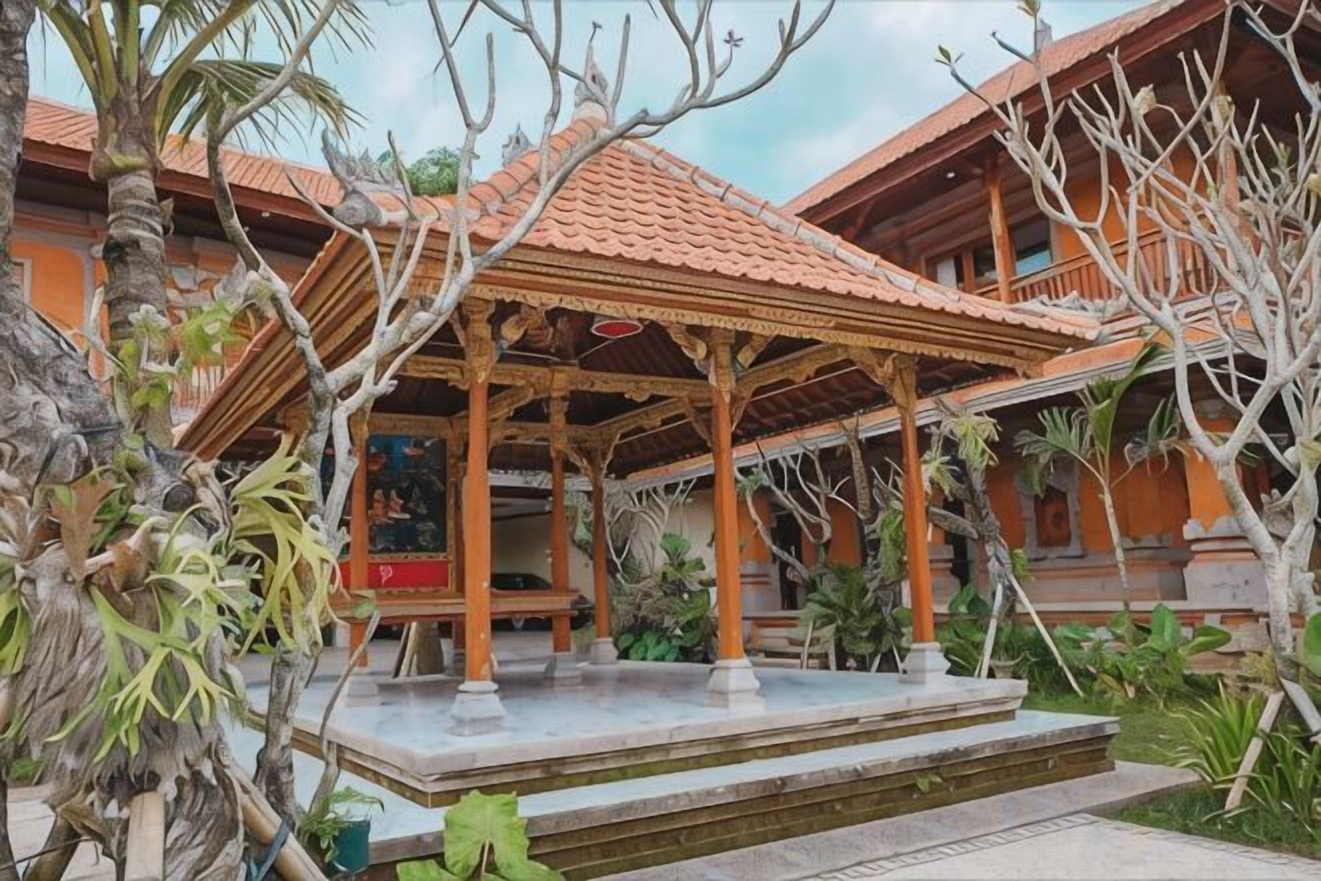En traditionel træpergola med et fliset tag, omgivet af frodig vegetation og en bygning med flisetag i baggrunden.