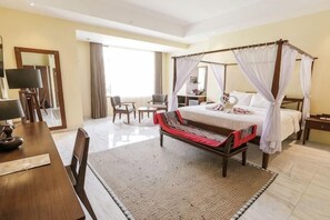 Una cama con dosel y cuatro columnas con un edred ón rojo y blanco, una mesita de noche de madera, una mesilla con lámpara y un ventanal con cortinas.