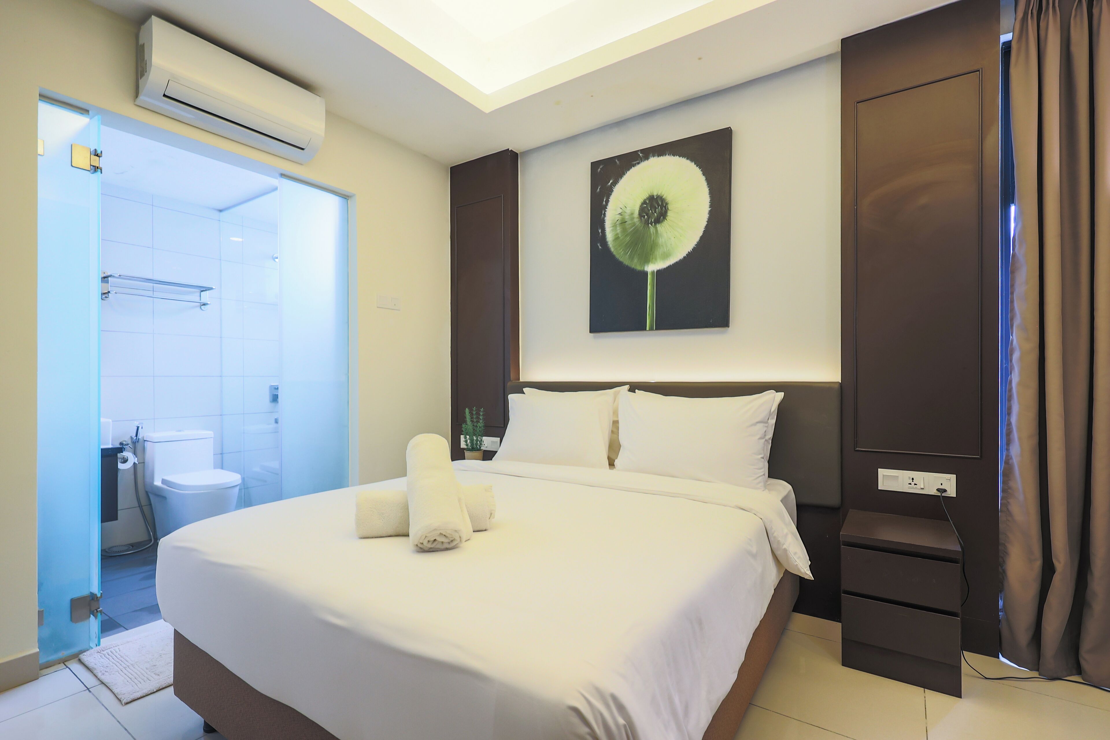 A hotel room with a bed, a nightstand, a wall-mounted air conditioner, a painting of a dandelion, and a bathroom visible through an open door.