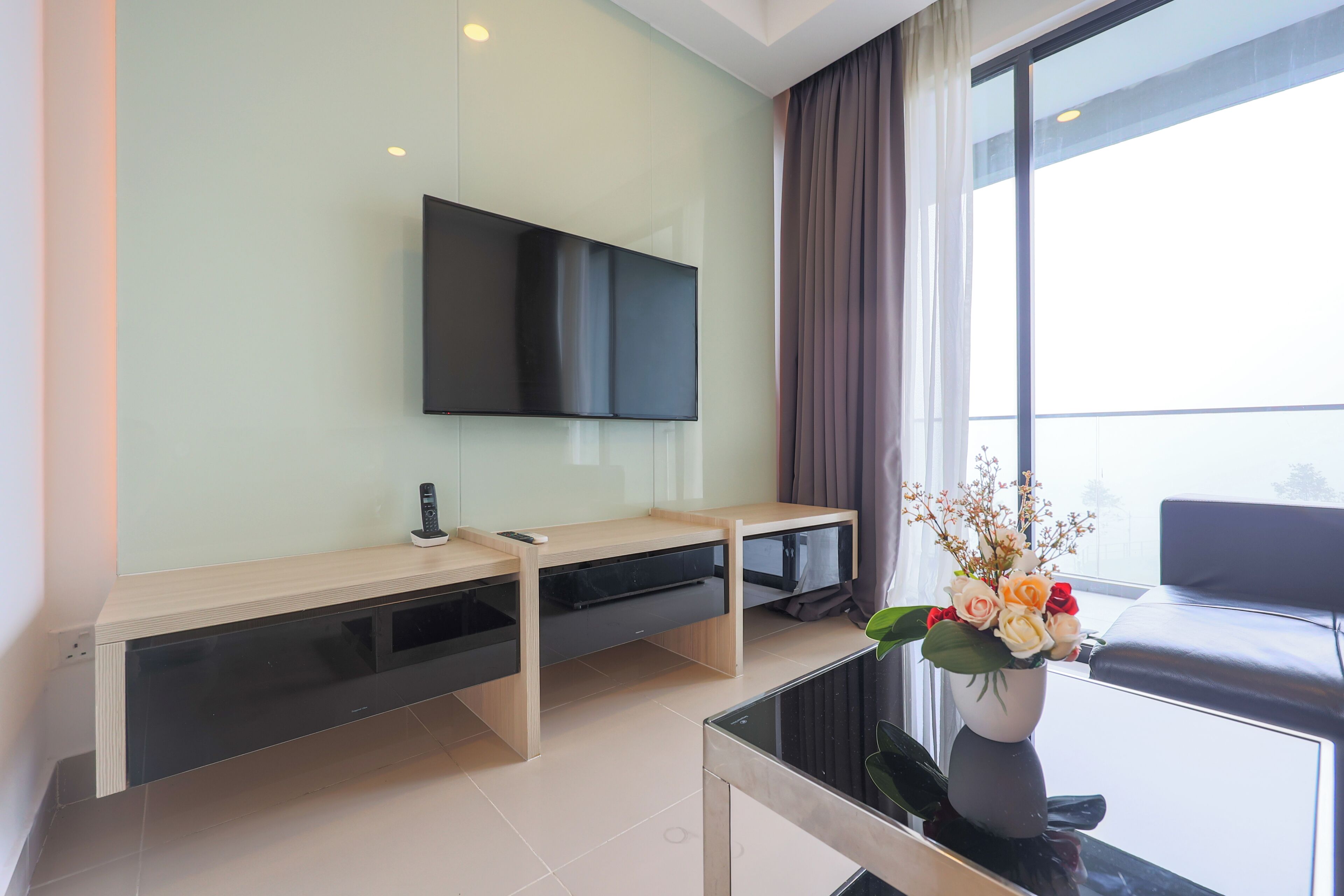 A modern living room with a flat-screen TV mounted on the wall, a wooden entertainment unit below, a glass coffee table, and a vase with flowers.