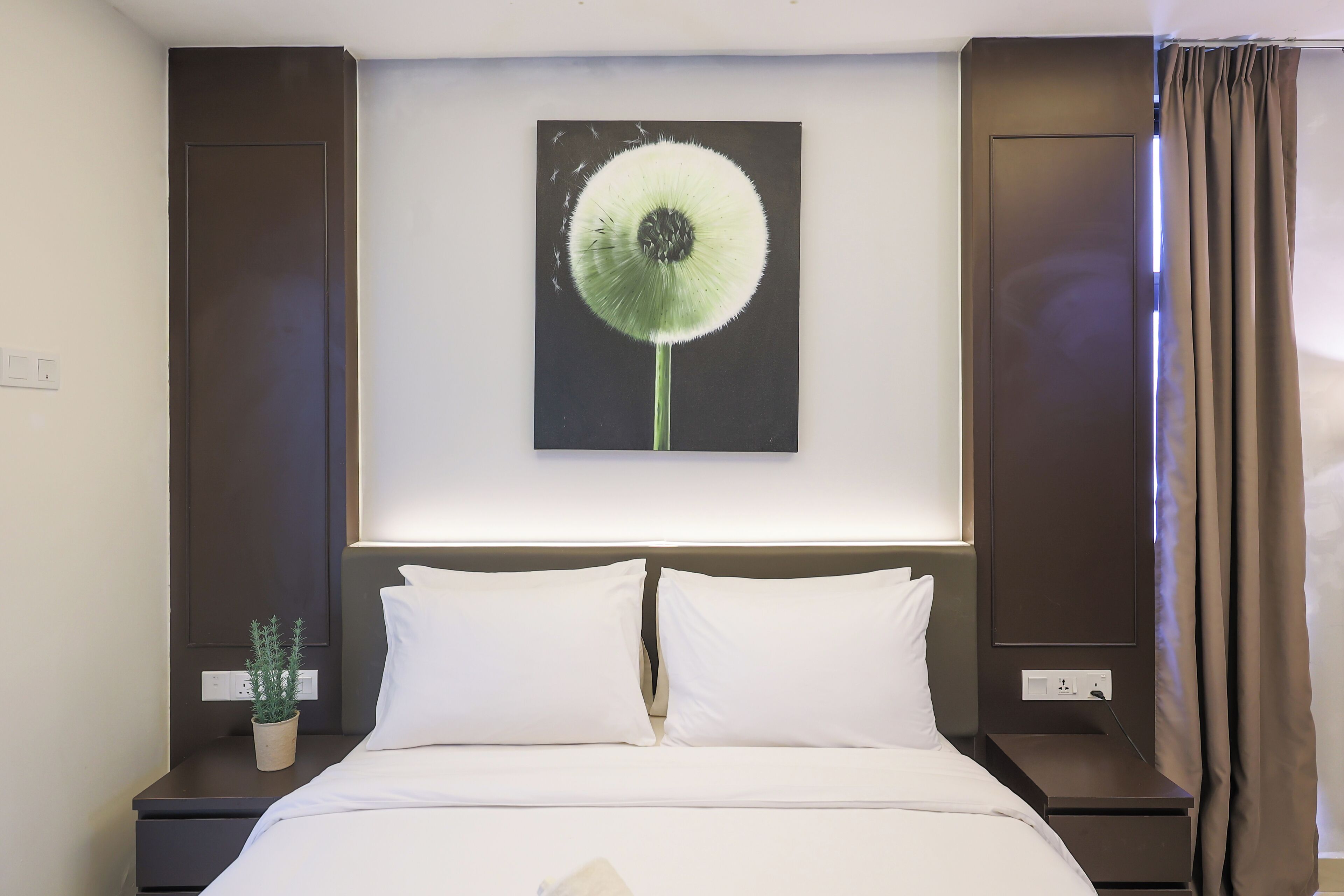 Quarto de hotel com uma cama grande, duas mesas de cabeceira, uma planta em vaso e uma obra de arte na parede retratando um dente-de-leão.
