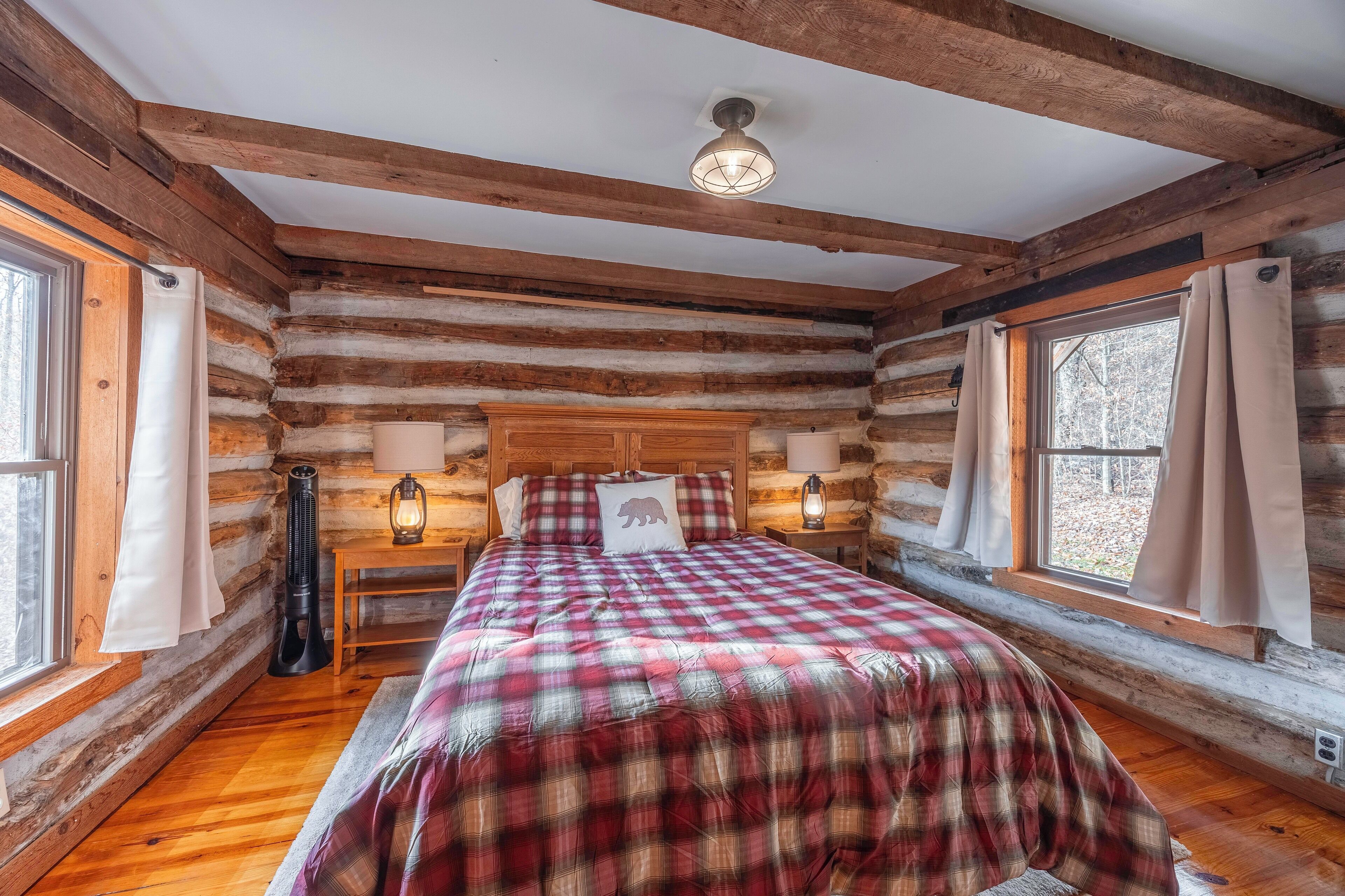A log cabin bedroom with a bed, two bedside tables with lamps, a window with curtains, and a ceiling fan.