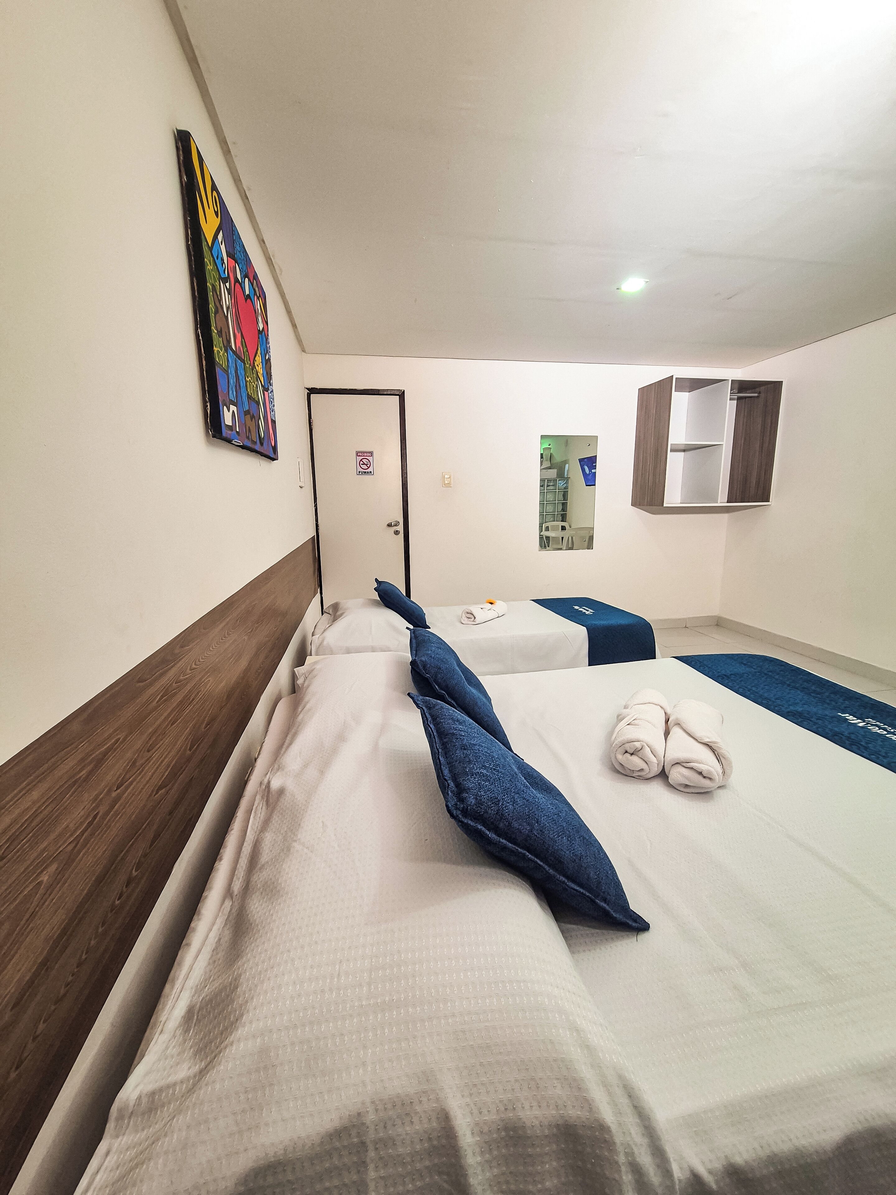 Une chambre d’hôtel avec deux lits, une porte sur laquelle est apposée une pancarte « Interdiction de fumer », une œuvre d’art colorée accrochée au mur et un miroir sur le mur.