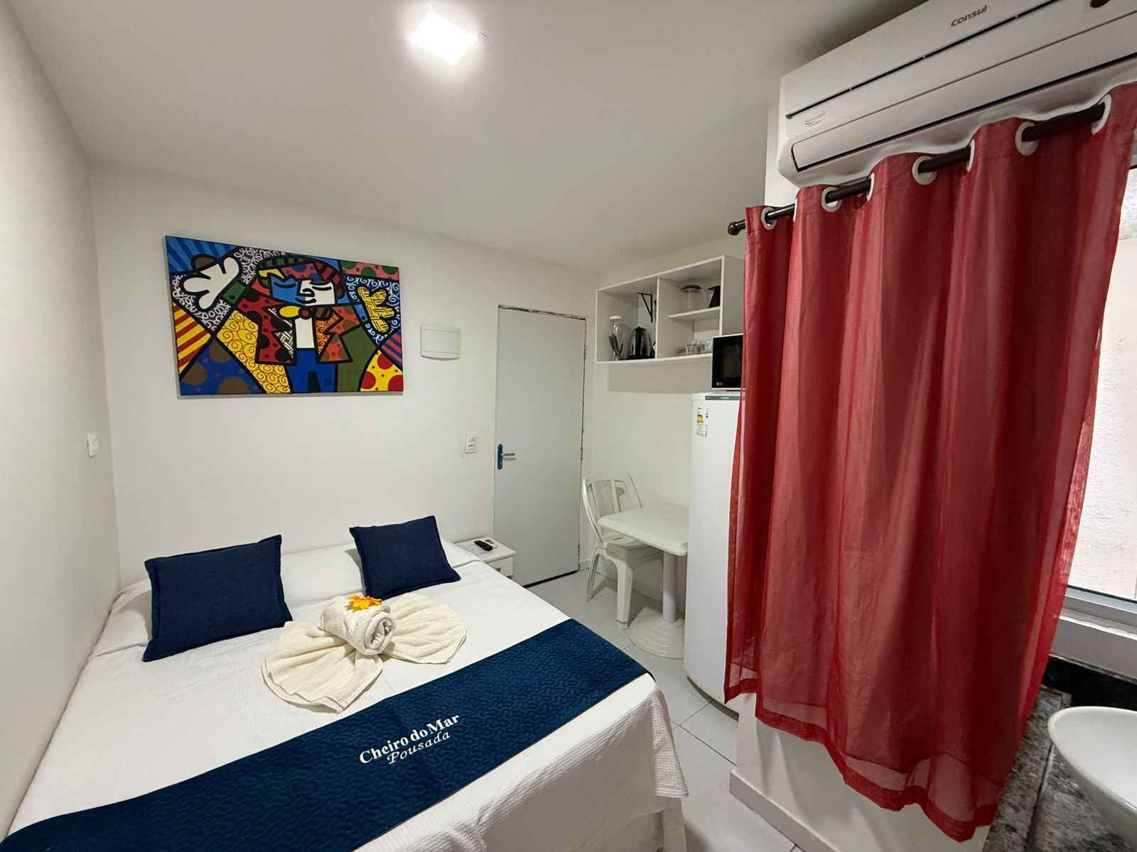Ein Hotelzimmer mit Bett, Schreibtisch, Stuhl, einem roten Vorhang und einem farbenfrohen Kunstwerk an der Wand.