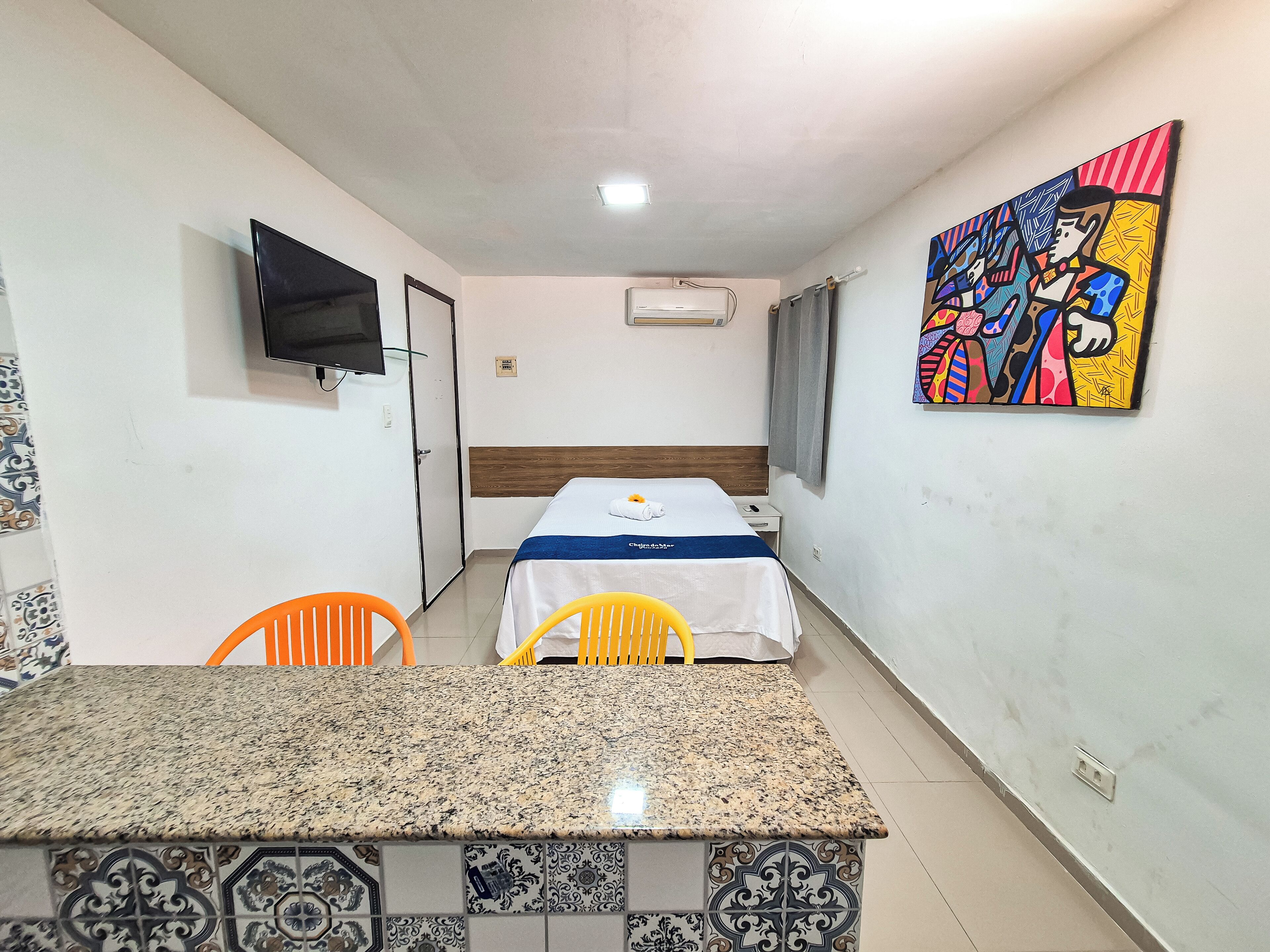 A hotel room with a bed, a TV, a painting, and a dining area with chairs and a table.