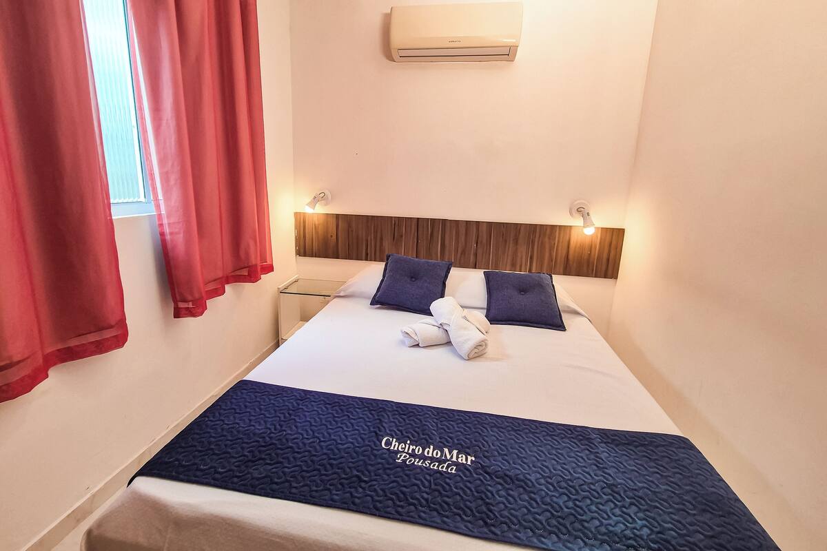 A hotel room with a bed, red curtains, an air conditioning unit, and a headboard with wall-mounted lights.