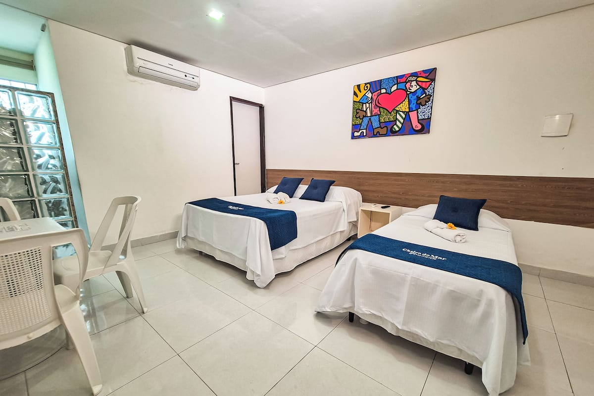 A hotel room with two beds, a small table, and a chair. There is a painting on the wall and an air conditioning unit above the door.
