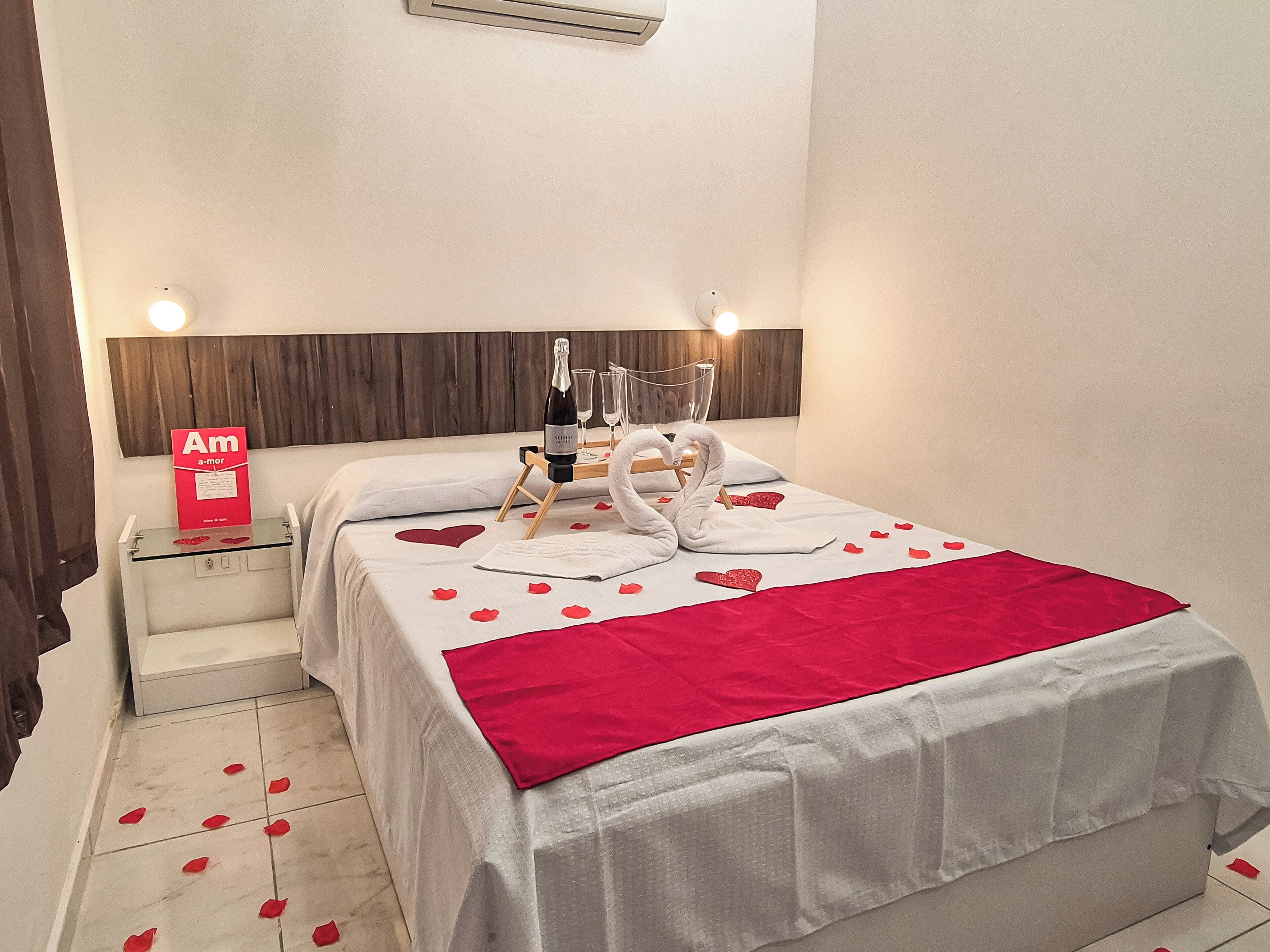 Una habitación de hotel con una cama bien hecha, una mesita de noche con una botella de champán, copas y un cartel que dice 'Am a-mor'.
