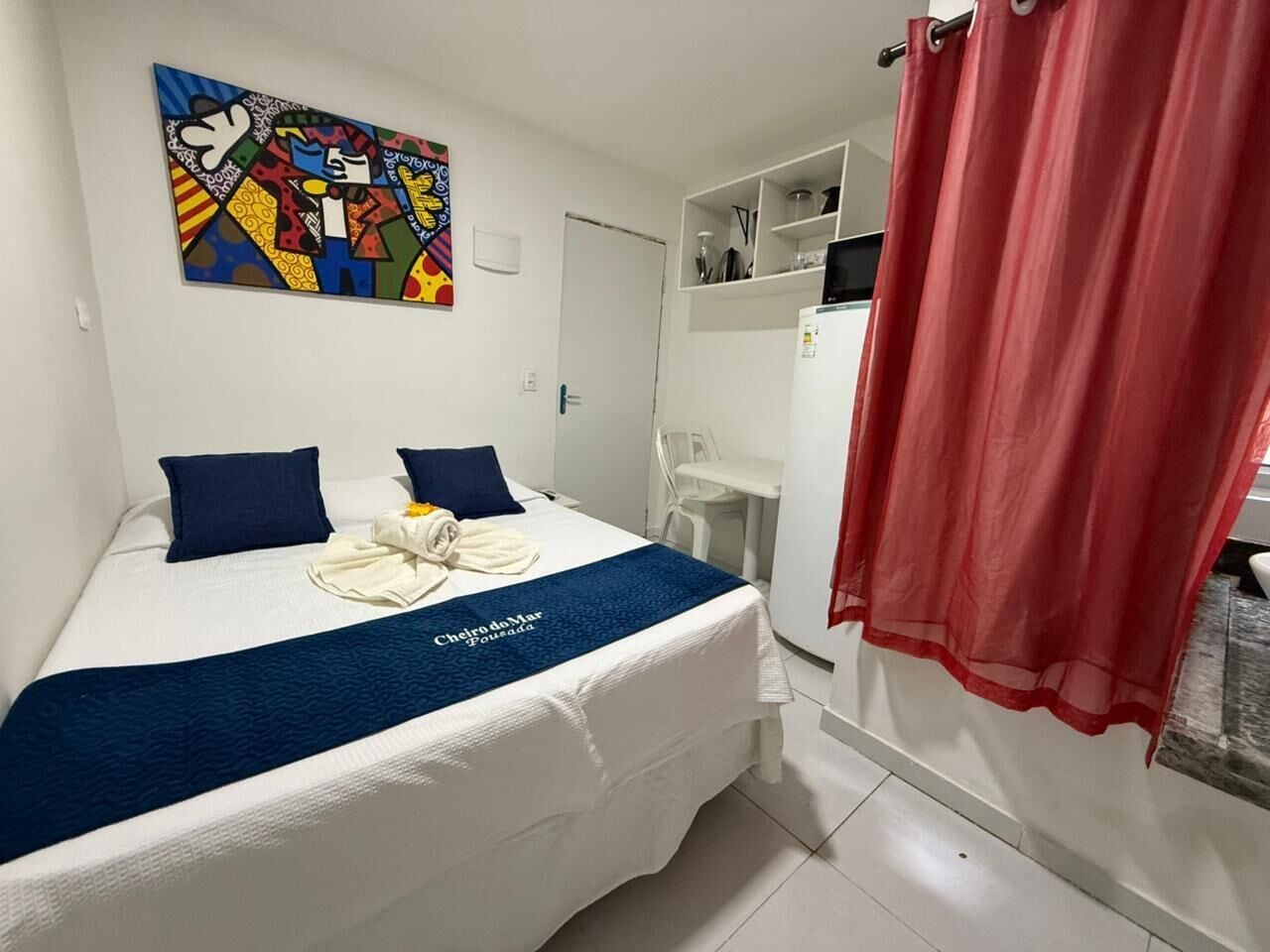 A hotel room with a bed, a painting on the wall, a red curtain, and a small table with a chair.