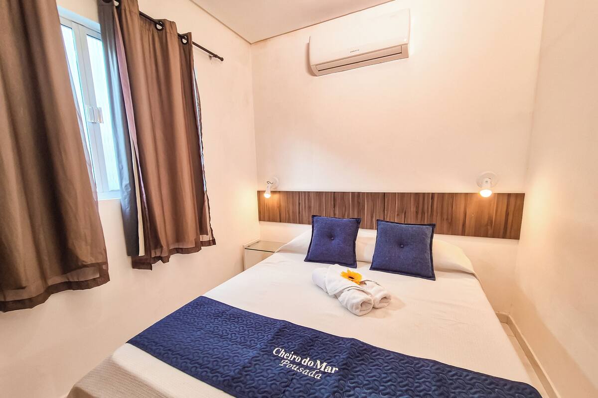 A hotel room with a bed, a nightstand, a wall-mounted air conditioner, and a window with curtains.