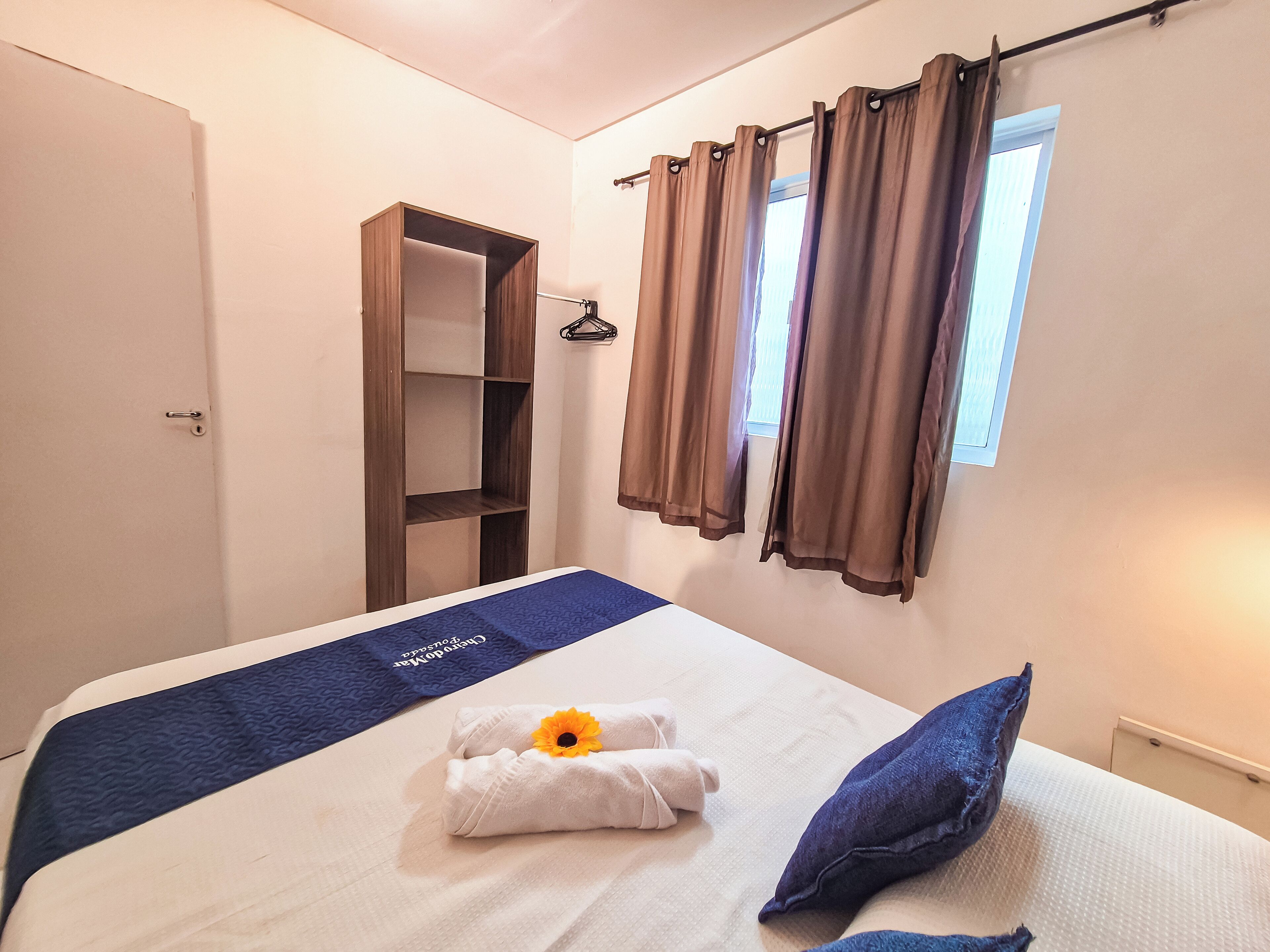 Una cama bien hecha con colcha azul, una toalla con un girasol y una almohada azul. Hay un armario con espejo y un estante, y una ventana con cortinas.