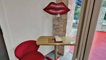 Una habitación pequeña con una mesa de madera, una silla roja, una lámpara decorativa con forma de labios rojos, y un cartel que dice 'Be A Doll Guest Book'.