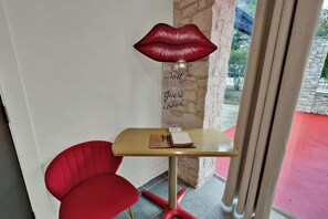 A small room with a wooden table, a red chair, a decorative red lips lamp, and a sign that reads "Be A Doll Guest Book".