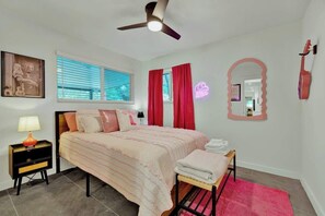 Ein Schlafzimmer mit einem Bett, einem Nachttisch, einem Spiegel, einem Deckenventilator und einem Fenster mit Jalousien.