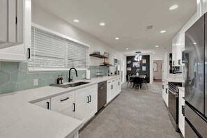 A modern kitchen with white cabinetry, a central island, and a dining area with a table and chairs. - Walk to Whitewater Amphitheater-GameRoom & Firepit (Canyon Lake)