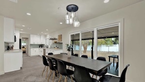 Une cuisine moderne avec des meubles blancs, un coin repas avec une longue table et des chaises noires, et une grande porte-fenêtre coulissante donnant sur un espace extérieur.
