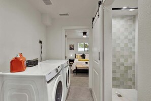 A laundry area with a washing machine and dryer, a small table with a bottle and basket, and a bedroom visible in the background. - Walk to Whitewater Amphitheater-GameRoom & Firepit (Canyon Lake)