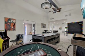 A spacious room with a pool table, a dartboard, and a blackjack table. There is a chalkboard with writing on it and a ceiling fan.