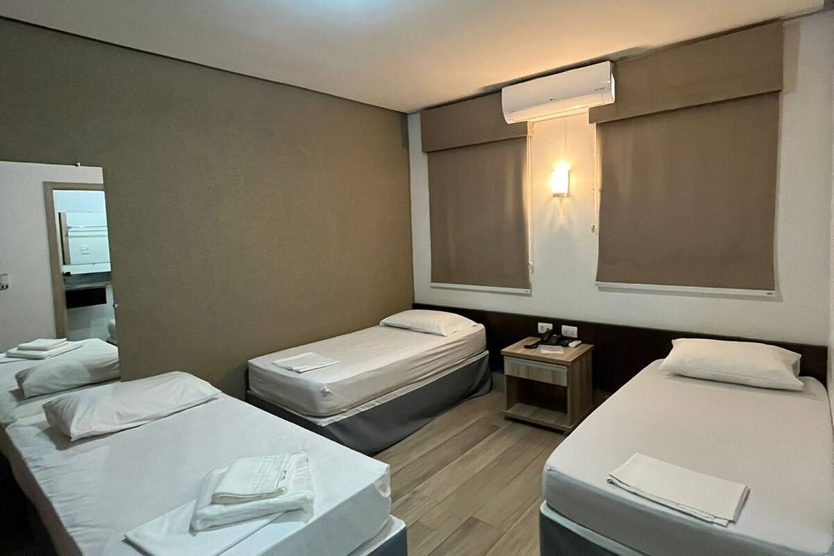 Une chambre d’hôtel avec deux lits simples, une petite table de chevet avec une lampe, une grande fenêtre avec des rideaux et un luminaire au plafond.