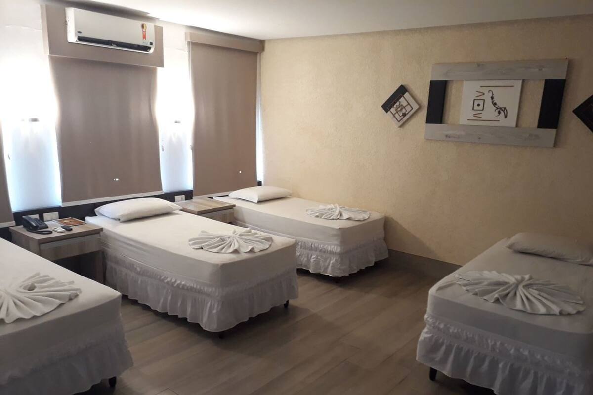 Une chambre d’hôtel avec trois lits simples, chacun agrémenté d’une serviette soigneusement pliée. On y trouve une table de chevet avec un téléphone et un livre, ainsi qu’un climatiseur fixé au mur.